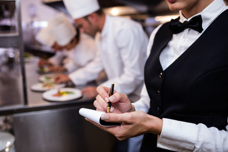 Close-up of waitress with notepad in commercial kitchen and chefs preparing food in backgroundの写真素材