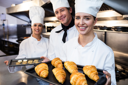 Three chefs in kitchen holding a tray of baked croissant and cookies in commercial kitchen