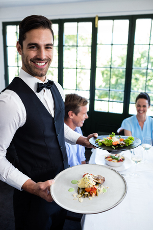 Portrait of waiter standing with meal next to customers in a restaurantの写真素材