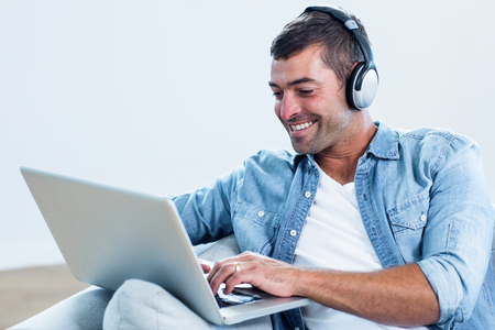 Young man listening to music while using laptop in living roomの写真素材