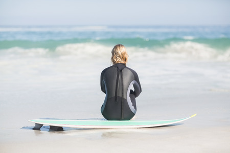 Rear view of woman in wetsuit sitting with surfboard on the beach on a sunny dayの写真素材