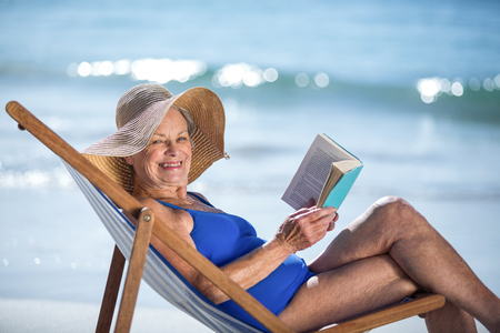 Pretty mature woman reading a book lying on deck chair on the beachの写真素材