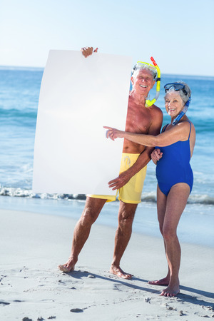 Senior couple with beach equipment holding a white poster at the beachの写真素材