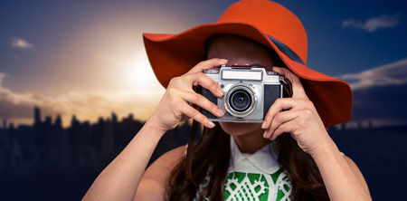Woman with hat taking photograph with camera against picture of city by sunriseの写真素材