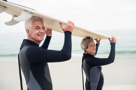 Portrait of senior couple in wetsuit carrying surfboard over head on the beachの写真素材