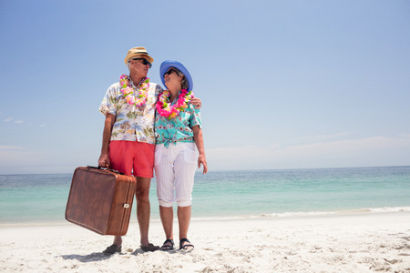 Happy senior couple wearing a garland and holding suitcase on the beachの写真素材