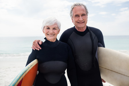 Portrait of senior couple in wetsuit holding surfboard on beach on a sunny dayの写真素材