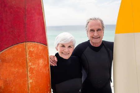 Portrait of senior couple with surfboard standing on the beach on a sunny dayの写真素材