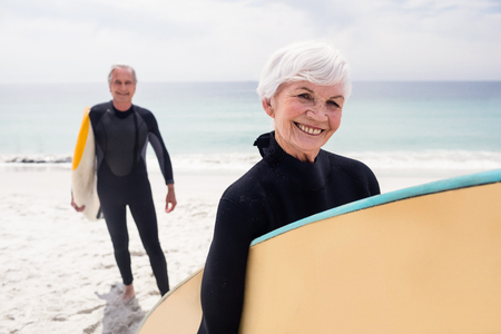 Portrait of senior couple in wetsuit holding surfboard on beach on a sunny dayの写真素材