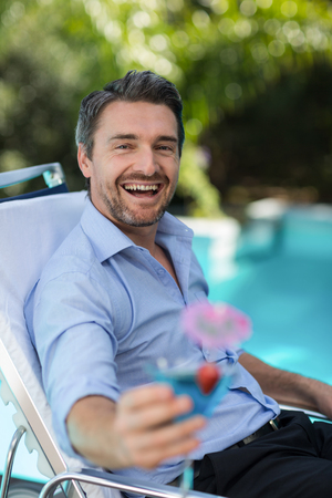 Smart man holding martini glass while relaxing in sun lounger near poolの写真素材