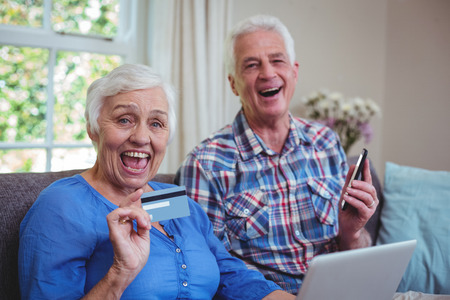 Portrait of cheerful senior couple with credit card and technology at homeの写真素材