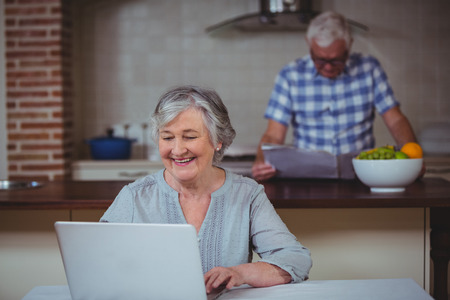 Happy senior woman using laptop with husband in background at kitchen