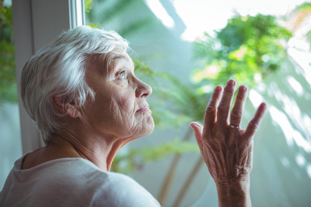 Thoughtful senior woman looking through house window at home