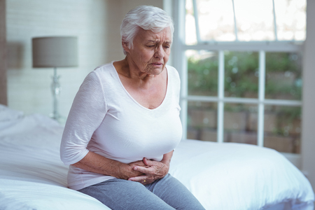Senior woman suffering from stomach pain while sitting on bedの写真素材
