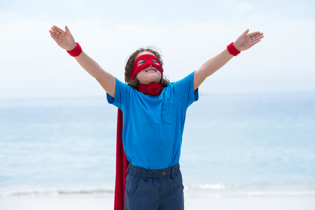Smiling boy in superhero costume standing with arms outstretched at sea shoreの写真素材