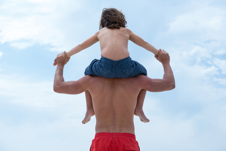 Rear view of father holding son hands while carrying him against skyの写真素材