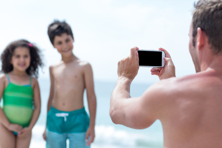 Father photographing smiling brother and sister at beachの写真素材