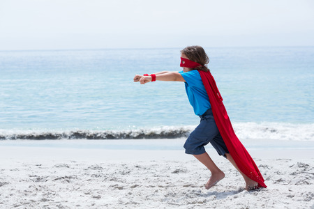 Full length of boy in superhero costume running with clenched fist at sea shoreの写真素材