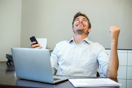 Smiling happy businessman holding mobile phone in officeの写真素材
