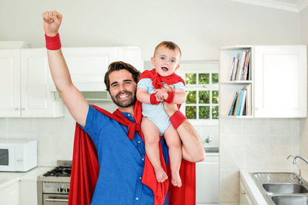 Happy father in superhero costume lifting son in kitchenの写真素材