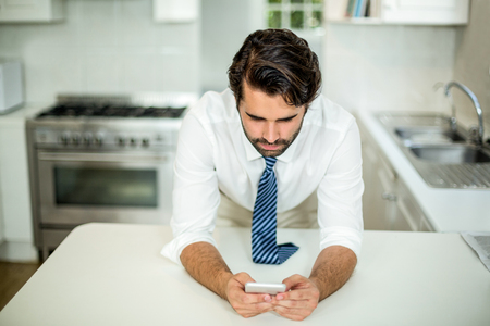 Businessman using cellphone at table in kitchen at homeの写真素材