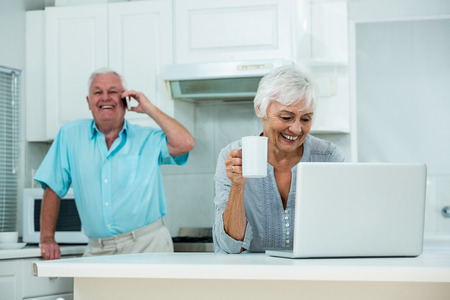 Happy woman using laptop while senior man talking on phone at homeの写真素材