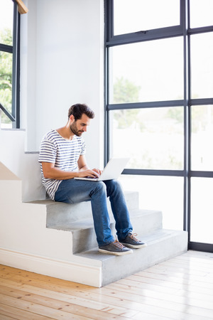 Happy man sitting on steps using laptop at homeの写真素材