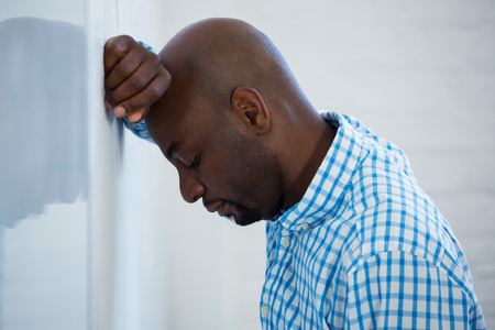 Upset man with eyes closed and hand on the forehead leaning against a wall in officeの写真素材