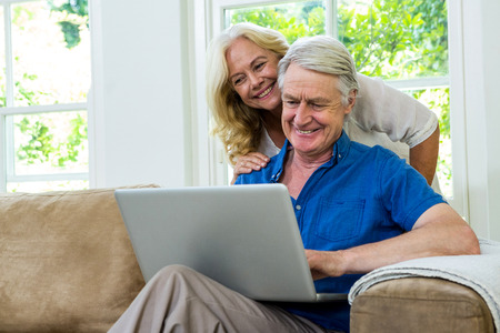 Happy senior couple using laptop while sitting on sofa against window at homeの写真素材