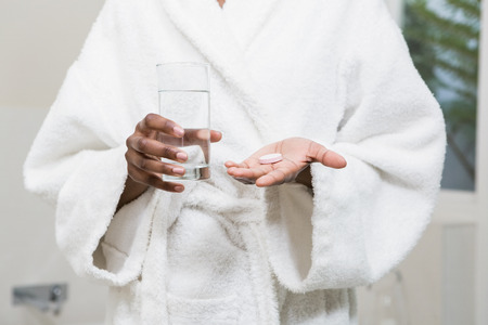 Beautiful woman holding pill and glass of water in the bathroomの写真素材