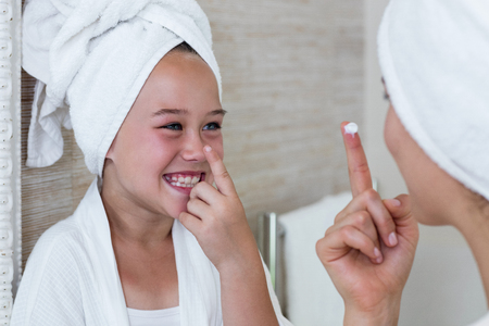 Smiling mother and daughter applying moisturizer on nose in bathroomの写真素材