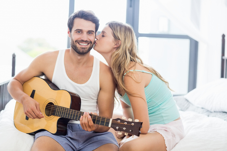 Man playing guitar while woman kissing him on cheek in the bedroomの写真素材