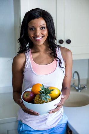 Portrait of young woman standing with bowl of fruits in kitchenの写真素材