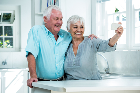 Cheerful retired couple taking selfie while standing in kitchenの写真素材
