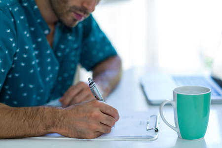 Mid-section of man writing on clipboard with coffee mug on table at homeの写真素材