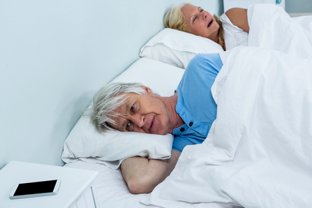 Thoughtful senior man relaxing on bed with phone on table at homeの写真素材
