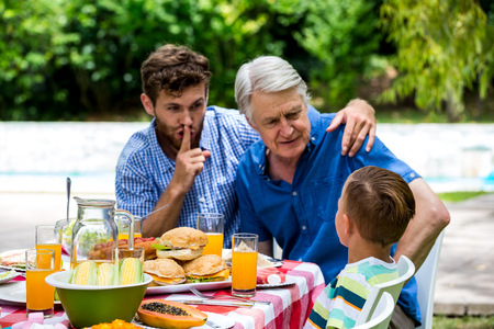 Man gesturing silence while son talking with grandfather at dinning table in lawnの写真素材