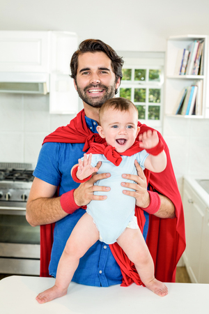 Happy father and son in superhero costume by table in kitchen at homeの写真素材