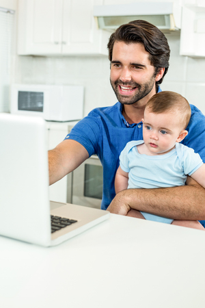 Cheerful man using laptop while sitting with son at table in kitchenの写真素材