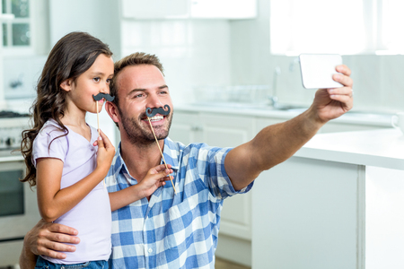 Father taking selfie with daughter holding artificial mustache stick at homeの写真素材