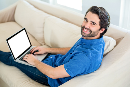 Portrait of happy handsome man using laptop while resting on sofa at homeの写真素材