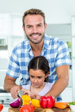 Father smiling while assisting daughter in cutting vegetables at homeの写真素材