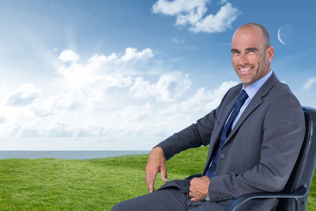 Portrait of confident businessman sitting on chair against green field under blue skyの写真素材