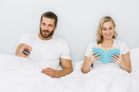 Man watching television while woman reading book at bedroomの写真素材
