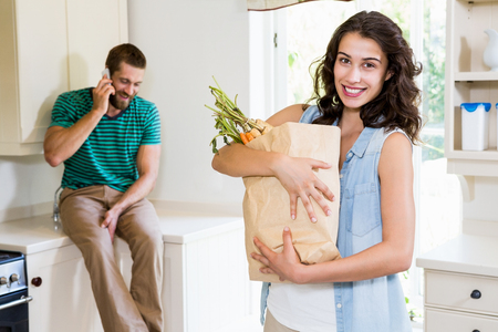 Woman holding groceries while man talking on mobile phone in kitchen at homeの写真素材