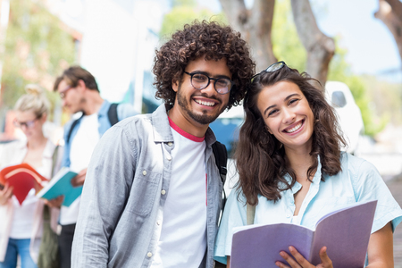 Portrait of students holding book while their friends interacting in the backgroundの写真素材