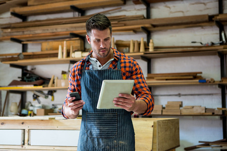 Carpenter reading a manual on the one hand and the other is taking his phone in a dusty workshop