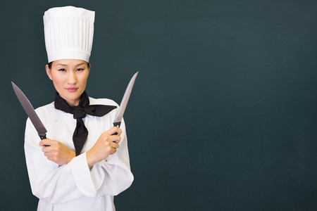 Confident female cook holding knives in kitchen against chalkboardの写真素材