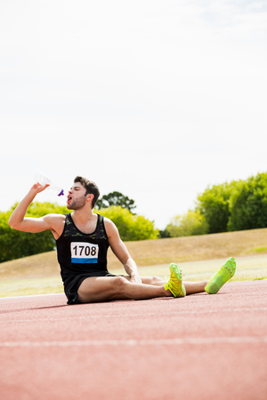 Tired athlete sitting on the running track and drinking water on a sunny dayの写真素材