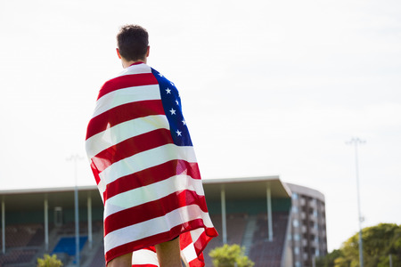 Rear view of athlete with american flag wrapped around his body in stadiumの写真素材